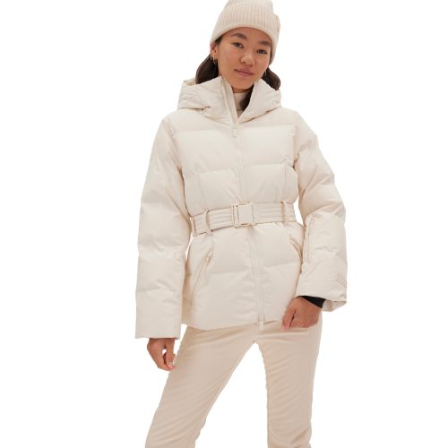 A person stands against a white background, wearing the Halfdays Hedy Puffer Jacket in cream, complete with a belt, matching pants, and a knitted beanie. The outfit's mountain performance features make it ideal for the slopes.