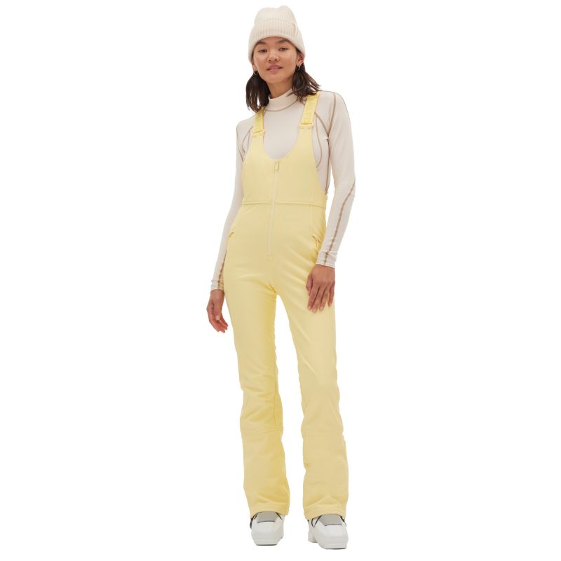 A person wearing the Isabel Soft Shell Bib Pant in a light yellow slim-fit style and a white beanie, standing and smiling against a plain background.